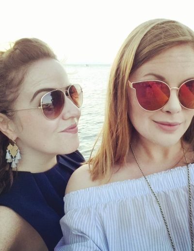 Alice y su mejor amiga sonriendo para una selfie frente al atardecer icónico de Santorini, con los edificios encalados de la isla de fondo