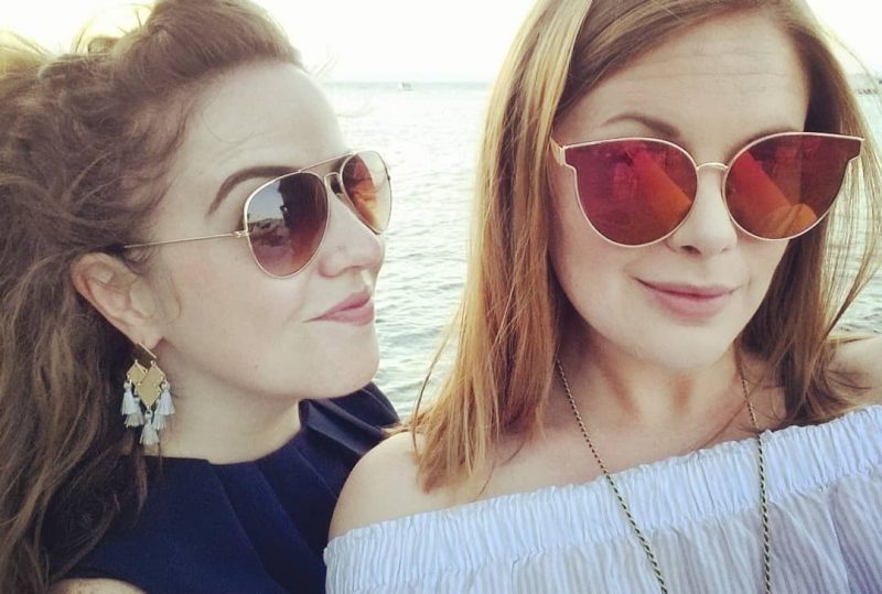 Alice y su mejor amiga sonriendo para una selfie frente al atardecer icónico de Santorini, con los edificios encalados de la isla de fondo