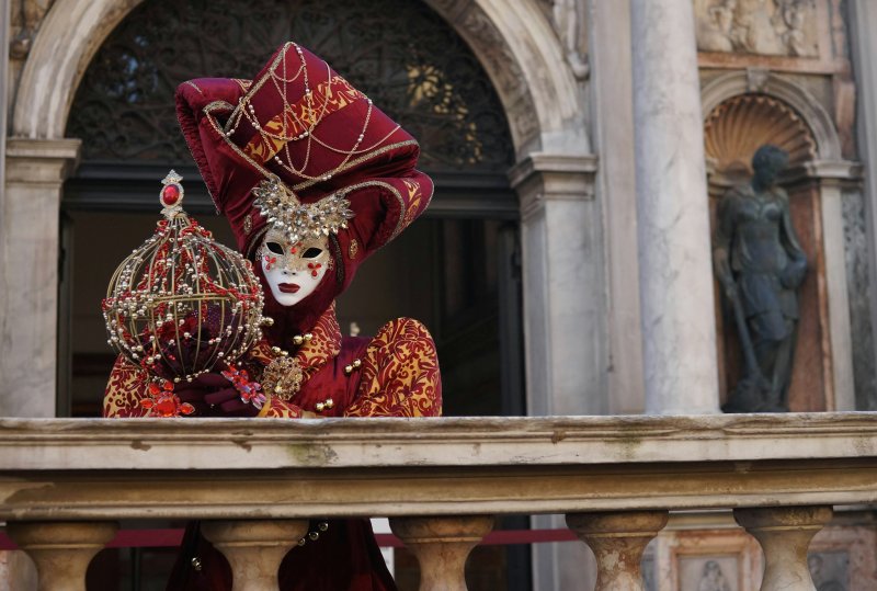 Person wearing ornate red and gold Venetian carnival mask with feathers standing on a balcony