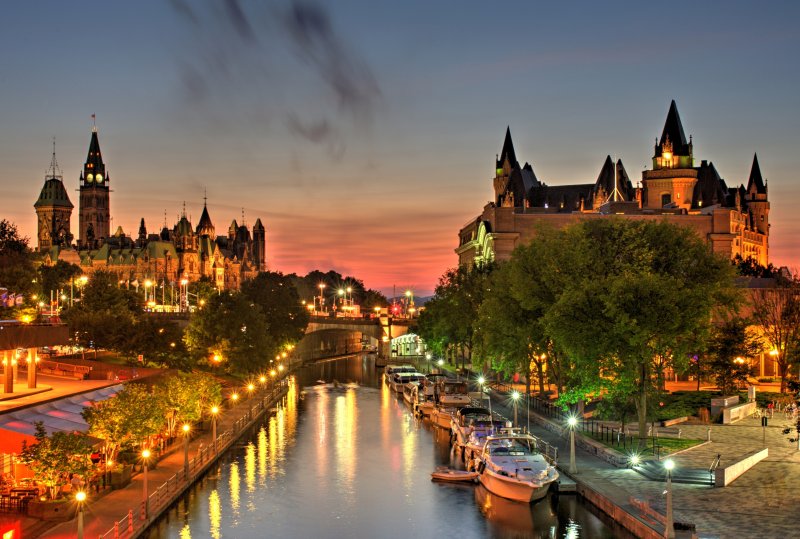 A beautiful warm orange-red glow fills the sky at dusk over Ottawa Canada during a July evening. A small wisp of low level clouds are visible. Parliament Hill, Chateau Laurier hotel and the Rideau Canal a UNSECO world heritage site are in the foreground.